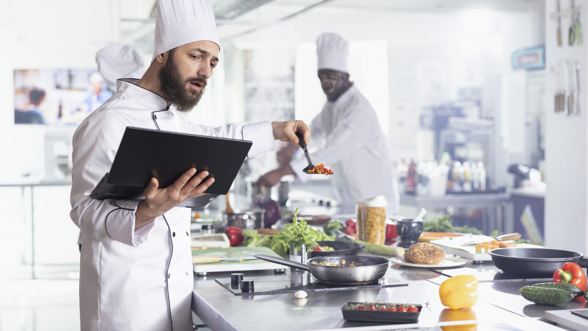 Focused on culinary research, male chef in a busy restaurant kitchen uses a laptop to find new recipe ideas while preparing ingredients. Man cooking gourmet food and plating delicious dishes.